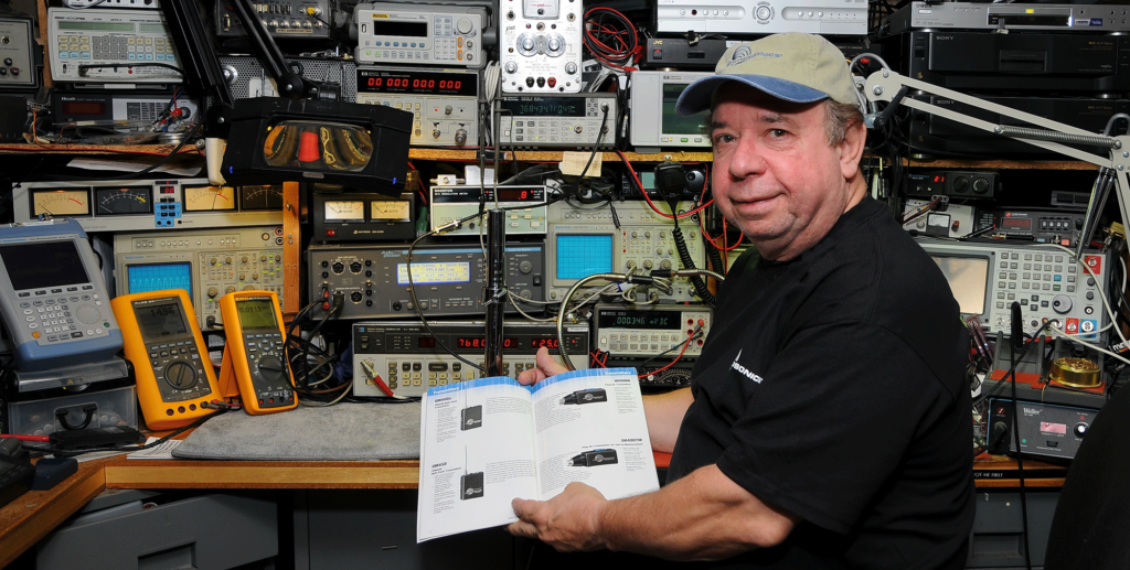 A man holding an electronic device in front of many other electronics.