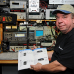 A man holding an electronic device in front of many other electronics.